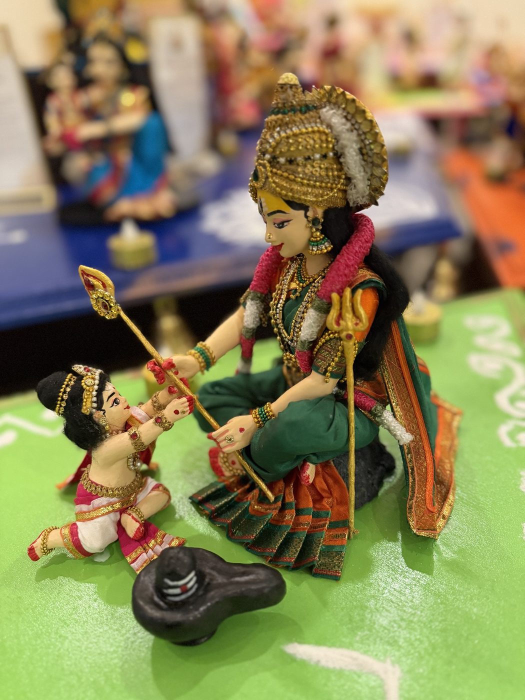 Golu doll of Parvati handing the Vel to little Murugan, who reaches up for it beside a Shiva lingam